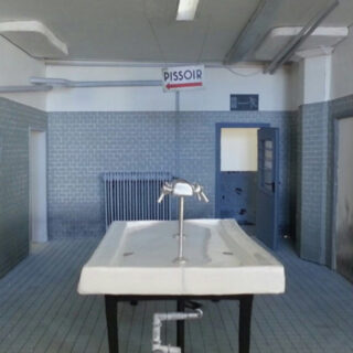 Ein gefliester Raum mit einem weißen Waschbecken in der Mitte, Rohren darunter, einem Heizkörper, einer offenen Tür und einem „PISSOIR“-Schild, das von der Decke hängt.