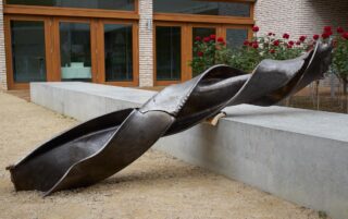 Eine große, verdrehte Metallskulptur steht auf einer Betonbank vor einem Gebäude mit Glastüren und roten Rosenbüschen.