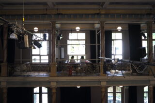 Zwei Personen sitzen an einem Schreibtisch im Balkonbereich eines Gebäudes, umgeben von Beleuchtungsgeräten und großen Fenstern, durch die natürliches Licht einfällt.