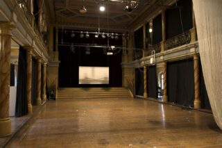 Ein leeres Theater mit Holzboden, Bühne, Projektionsleinwand, Deckenbeleuchtung und verzierten Säulen, von der Mitte des Zuschauerraums aus gesehen.