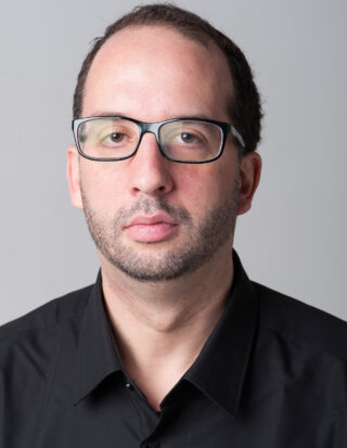 Ein Mann mit kurzen dunklen Haaren, Brille und kurzem Bart trägt ein schwarzes Hemd mit Kragen und blickt vor einem schlichten hellen Hintergrund direkt in die Kamera.