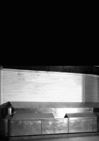 Vor einer weißen Wand mit Fensterläden und einem schwarzen Hintergrund steht eine leere Verkaufstheke aus Edelstahl.