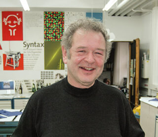 Ein Mann mit lockigem grauem Haar und einem dunklen Pullover lächelt in einem Designstudio, an der Wand hinter ihm hängen bunte Poster und Kunstwerke.