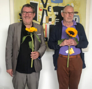 Zwei ältere Männer stehen nebeneinander im Haus, jeder hält eine Sonnenblume in der Hand, an der Wand hinter ihnen hängt ein farbenfrohes abstraktes Gemälde.