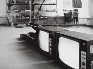 Zwei alte Fernseher und ein Plattenspieler stehen auf dem Boden eines Raumes, im Hintergrund stehen Metallregale, Bücher und ein Schreibtisch.