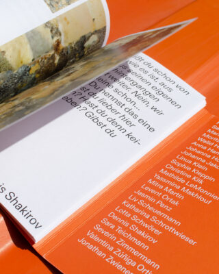 Ein aufgeschlagenes Buch auf orangefarbener Oberfläche zeigt eine Seite mit deutschem Text und eine weitere Seite mit Namen in weißer Schrift auf orangefarbenem Hintergrund.