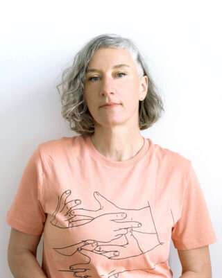 Vor einem schlichten weißen Hintergrund steht eine Person mit kurzen grauen Haaren und einem hellrosa Hemd mit einer Strichzeichnung von Händen darauf.