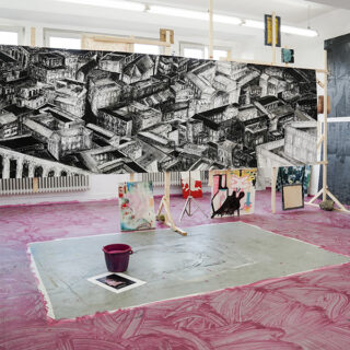 Ein Kunstatelier mit einem großen schwarz-weißen Stadtbild, verschiedenen abstrakten Kunstwerken drumherum und einem mit rosa Farbe bedeckten Boden mit einem lila Eimer.