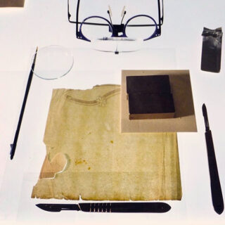 Eine Brille, eine Lupe, ein Pinsel, ein Notizblock, eine Schachtel, ein Metallskalpell und ein kleines Metallwerkzeug, angeordnet auf einer flachen Oberfläche.