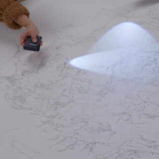 Eine Hand beleuchtet mit einer kleinen Taschenlampe ein großes Blatt Papier und zeichnet detaillierte Bleistiftskizzen nach.