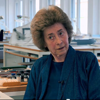 Eine ältere Frau mit kurzem, voluminösem Haar sitzt in einem Büro oder Labor. Sie trägt ein blaues Oberteil und eine Jacke, im Hintergrund sind verschiedene Geräte zu sehen.