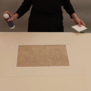 Eine Person in schwarzer Kleidung hält eine Sprühkleberdose und eine weiße Karte über einem rechteckigen Blatt Papier auf einem beigen Tisch.