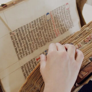 Eine Hand blättert vorsichtig die abgenutzten Seiten eines alten, illuminierten Manuskripts mit mittelalterlichem Text und farbigen Initialen um.