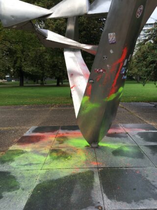 Eine silberne Metallskulptur mit Aufklebern und Spritzern aus roter und grüner Farbe steht auf einer gepflasterten Fläche in einer Parkanlage.
