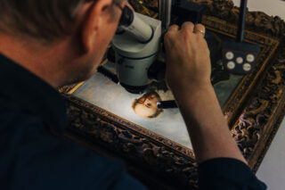 Eine Person untersucht ein gerahmtes Porträtgemälde durch ein Mikroskop und konzentriert sich auf das Gesicht im Kunstwerk.