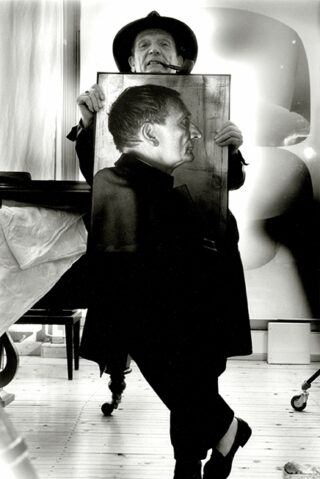 Ein älterer Mann mit Hut und Mantel und einer Pfeife im Mund steht im Haus und hält auf Brusthöhe ein gerahmtes Porträt von sich im Profil.