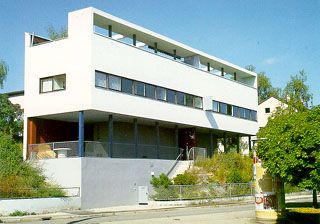 Ein modernistisches weißes rechteckiges Gebäude mit Flachdach, großen horizontalen Fenstern und einer erhöhten, von Säulen getragenen Hauptebene.