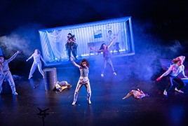 Eine Gruppe von Darstellern in dynamischen Posen auf einer rauchigen Bühne, mit einer beleuchteten Kulisse, die an einen Schiffscontainer erinnert, und verstreuten Requisiten.