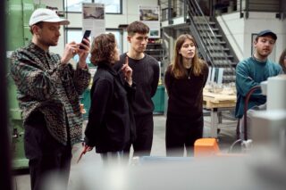 Eine Gruppe von fünf Personen steht in einem Gebäude in der Nähe von Maschinen; eine Person telefoniert, während die anderen aufmerksam zuschauen und zuhören.