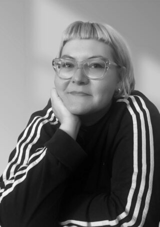 Person mit kurzen hellen Haaren und Brille, trägt ein dunkles Oberteil mit weißen Streifen an den Ärmeln, stützt das Kinn auf die Hand und blickt in die Kamera. Schwarz-Weiß-Foto.