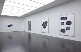 Eine moderne Kunstgalerie mit weißen Wänden, in der vier abstrakte, gerahmte Kunstwerke ausgestellt sind. Der Boden ist dunkel und reflektierend und der Raum wird durch Deckenpaneele beleuchtet.