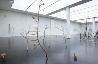 Kunstinstallation in einem Galerieraum mit kahlen Ästen, hängenden Objekten und Pilzskulpturen auf einem polierten schwarzen Boden und gerahmten Kunstwerken an weißen Wänden.