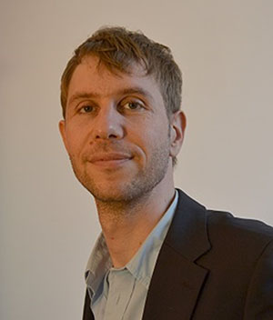 Ein Mann mit kurzen hellbraunen Haaren, einem dunklen Blazer und einem hellen Hemd posiert vor einem schlichten Hintergrund.