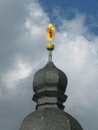 Ein Kirchturm mit einem grauen Schieferdach und einem kunstvollen Goldornament mit einer roten Figur an der Spitze vor einem bewölkten Himmel.
