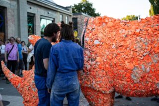Zwei Personen in blauen Overalls stehen bei einer Veranstaltung im Freien neben einer großen orangefarbenen Nashornskulptur, die aus verschiedenen zerbrochenen Keramikstücken besteht.