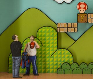 Zwei Figuren stehen vor einer Kulisse im Super-Mario-Stil mit grünen Hügeln, Fragezeichenblöcken und einem Power-Up-Pilz.