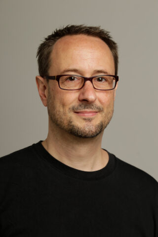 Ein Mann mit kurzen braunen Haaren, Brille und gestutztem Bart, der ein schwarzes Hemd trägt, steht vor einem schlichten hellgrauen Hintergrund.
