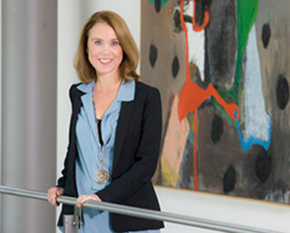 Eine Frau in einer blauen Bluse und einem schwarzen Blazer steht lächelnd an einem Geländer im Haus, hinter ihr hängt ein abstraktes Gemälde an der Wand.