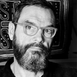 Schwarz-Weiß-Porträt eines Mannes mittleren Alters mit Bart und Brille, der in die Kamera blickt, mit einem gemusterten Textil im Hintergrund.