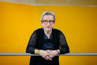 Eine Person mit kurzen hellen Haaren, roter Brille und schwarzem Outfit lehnt vor einem leuchtend gelben Hintergrund an einem Metallgeländer.