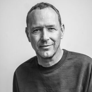 Schwarz-Weiß-Porträt eines Mannes mittleren Alters mit kurzen Haaren, einem leichten Lächeln und einem einfachen Sweatshirt mit Rundhalsausschnitt, der vor einem schlichten Hintergrund in die Kamera blickt.