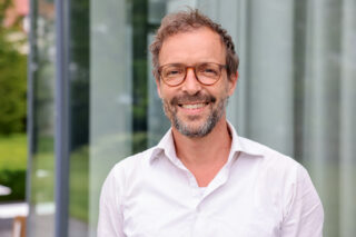 Ein Mann mit Brille und Bart, der ein weißes Hemd trägt, lächelt, während er draußen vor Glasfenstern steht.