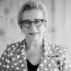 Eine Frau mit kurzen Haaren und Brille sowie einer gemusterten Jacke steht vor einem schlichten Hintergrund. Das Bild ist schwarz-weiß.