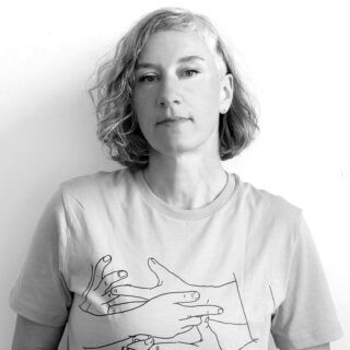 Eine Person mit kurzem, welligem Haar und einem T-Shirt mit einer Strichzeichnung von Händen darauf steht vor einem schlichten weißen Hintergrund.