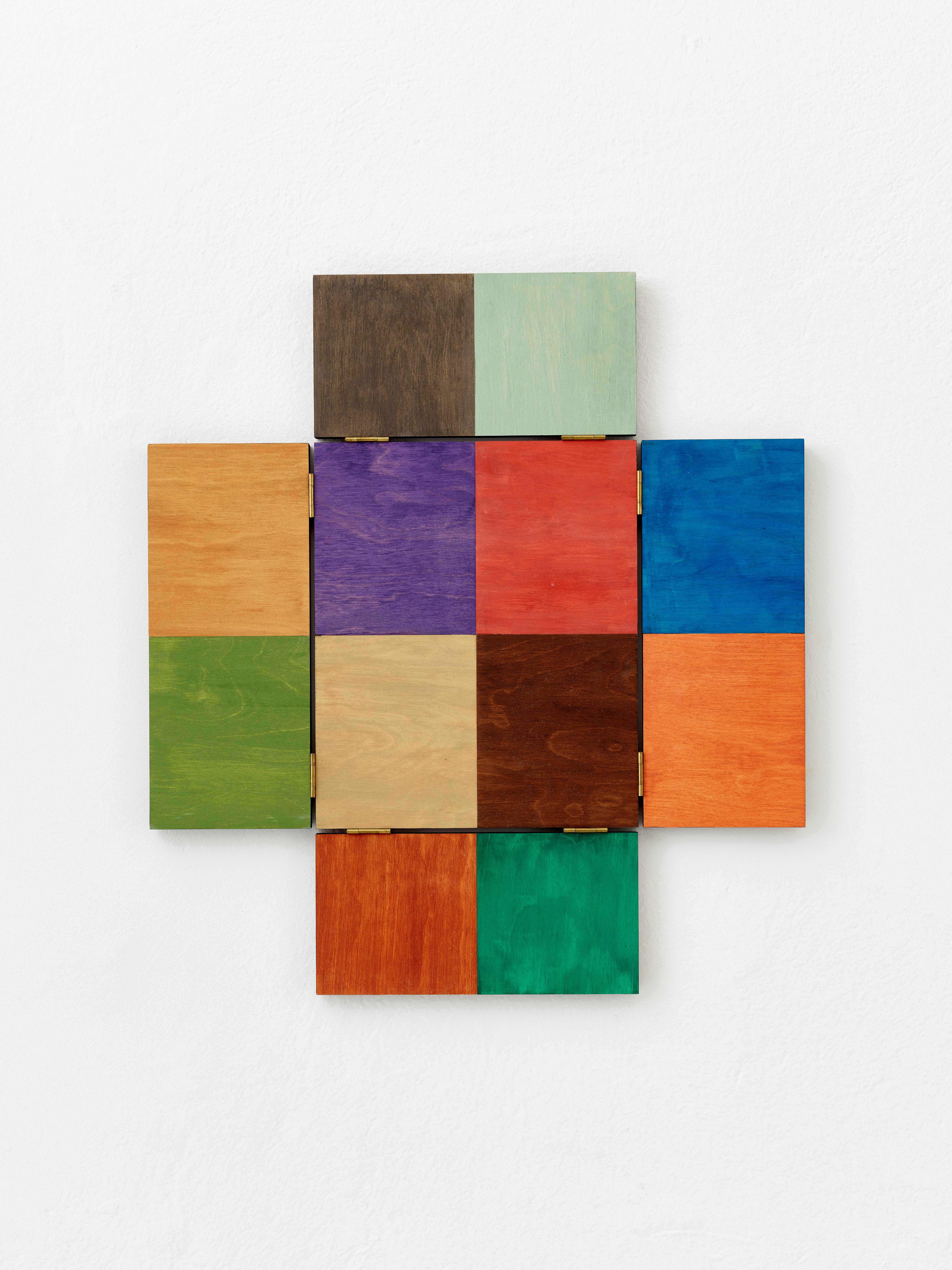 Ein Raster aus neun quadratischen Holztafeln, jede in einer anderen Farbe bemalt, kreuzförmig angeordnet und an einer weißen Wand angebracht.