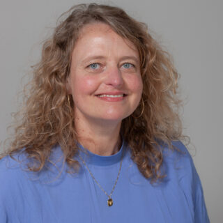 Frau mit lockigem blondem Haar, trägt ein blaues Oberteil und eine goldene Halskette und lächelt vor einem schlichten hellgrauen Hintergrund in die Kamera.