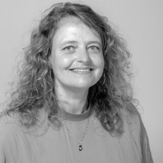 Eine Frau mit lockigem Haar, einem hellen Oberteil und einer Halskette, lächelt in einem Schwarz-Weiß-Porträt in die Kamera.