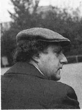 Ein Mann mit Schiebermütze und Mantel ist im Profil zu sehen, wie er im Freien sitzt, im Hintergrund sind unscharfe Bäume zu sehen.