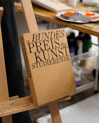 Ein Buch mit dem Titel „26. Bundespreis für Kunststudierende“ liegt auf einer hölzernen Staffelei in einem Kunstatelier, im Hintergrund sind Künstlerutensilien und eine Farbpalette zu sehen.