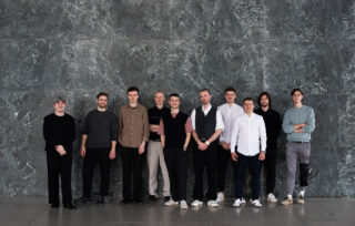 Zehn Männer stehen in einer Reihe vor einer grauen Marmorwand und blicken in die Kamera. Sie tragen legere und halbformelle Kleidung.