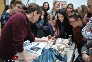 Eine Gruppe von Menschen beobachtet eine Frau, die auf einem Tisch die Restaurierung einer beschädigten Skulptur vorführt. In der Nähe befinden sich Referenzbilder und -materialien.