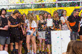 Eine Gruppe von Menschen steht inmitten von fallendem Konfetti auf einer Bühne vor einem leuchtend gelben Hintergrund. Einige halten ein Zertifikat oder Dokument in den Händen.