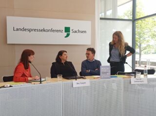 Vier Personen sitzen an einem Konferenztisch vor einem Schild mit der Aufschrift „Landespressekonferenz Sachsen“ und diskutieren. Auf dem Tisch sind Namensschilder und ein Buch zu sehen.