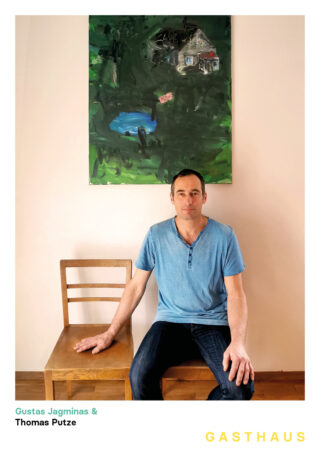 Ein Mann in einem blauen Hemd sitzt auf einem Holzstuhl neben einem anderen leeren Stuhl. An der Wand hinter ihm hängt ein Gemälde mit einem Haus und viel Grün.