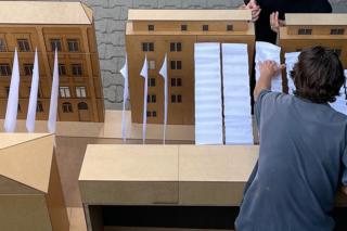 Eine Person ordnet weiße Papierblätter auf einem Architekturmodell von Gebäuden aus Pappe an, wobei die Hände einer anderen Person im Rahmen sichtbar sind.