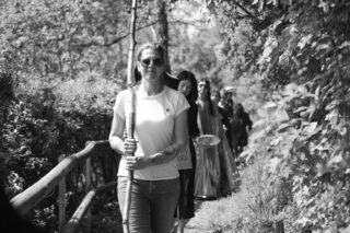 Eine Gruppe von Menschen geht einen Waldweg entlang, angeführt von einer Frau mit Sonnenbrille und einem Stab in der Hand. Die Szene ist in Schwarzweiß.